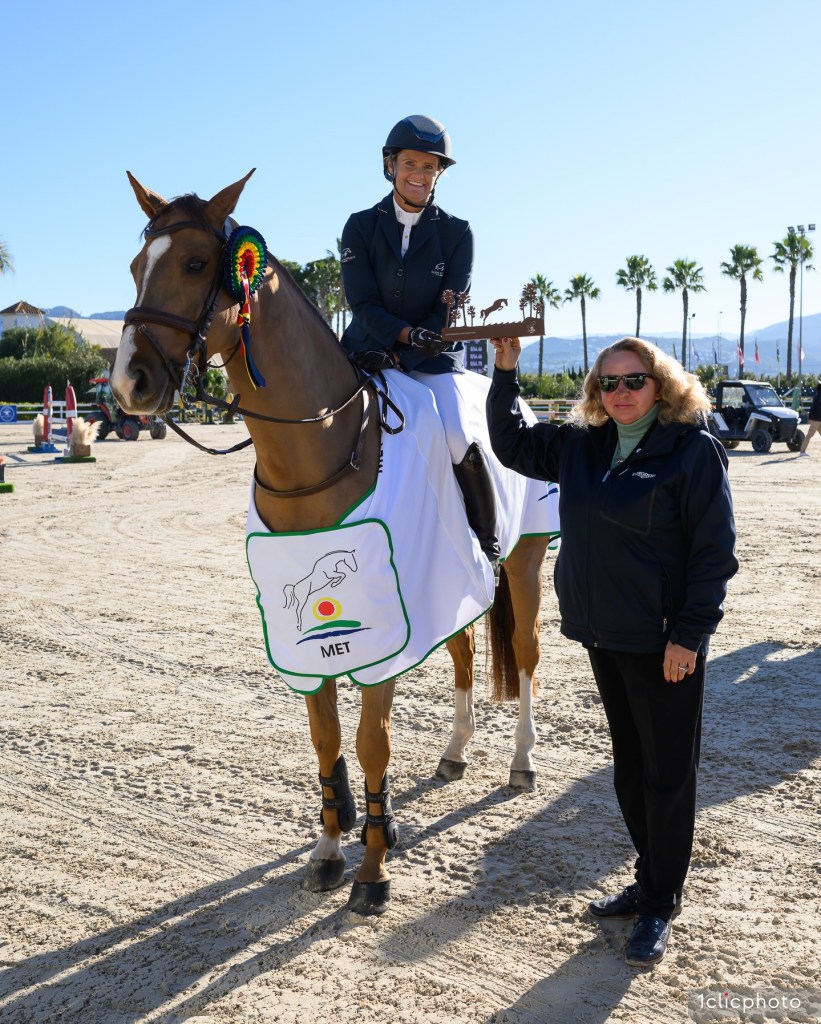 A smiling female rider in competition attire holds a trophy atop her horse, while another woman beside her celebrates at the Mediterranean Equestrian Tour in Spain.