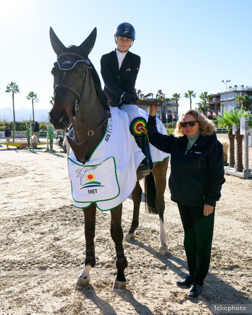 A rider in a black helmet and competition attire sits on a horse adorned with a white blanket that has a logo, while a woman in a black jacket stands next to them, both enjoying a moment of victory at the Mediterranean Equestrian Tour.