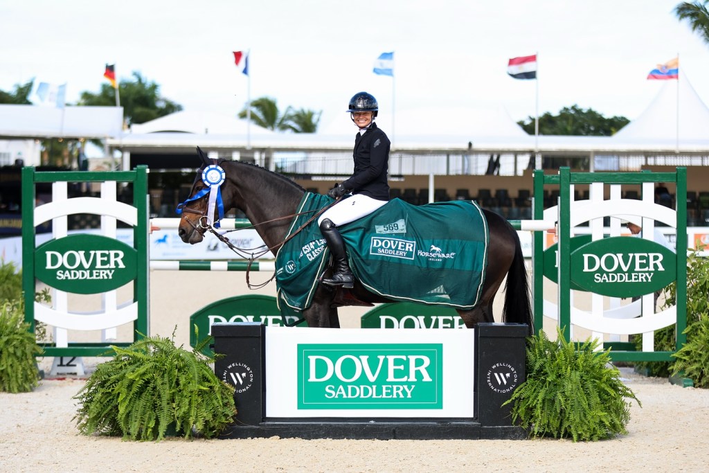A rider in competition attire and a horse wearing a decorative blanket stands proudly in front of a sign for Dover Saddlery, with flags in the background.