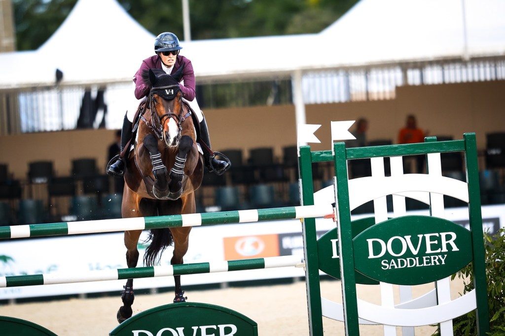 A rider in a helmet and purple jacket jumping over a hurdle with a 'Dover Saddlery' sign in the foreground during a horse jumping event.